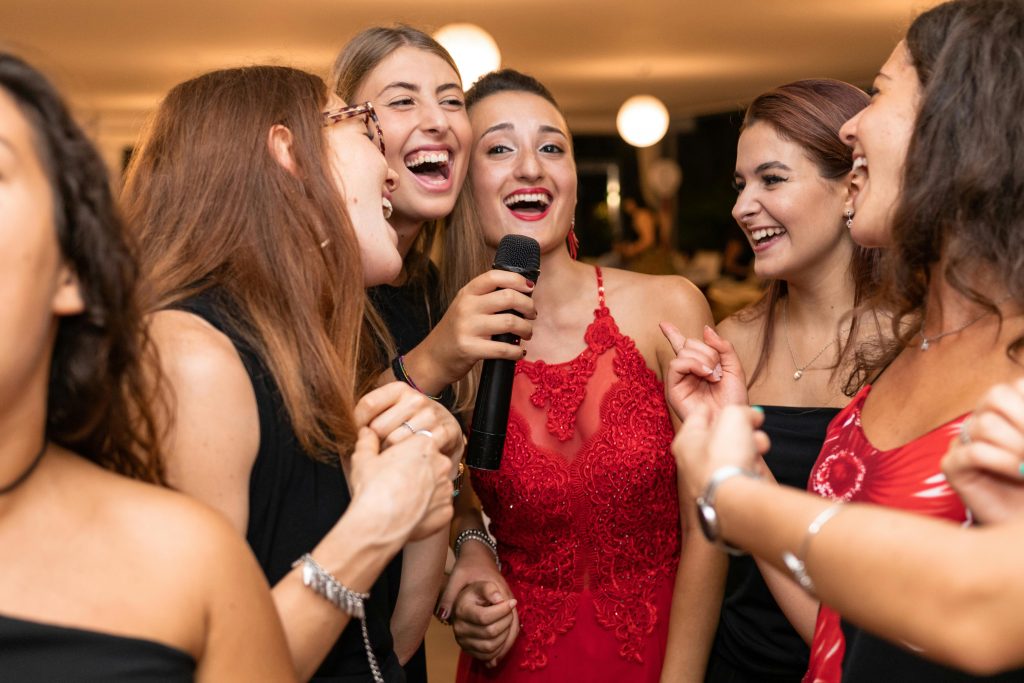 Group of cheerful ethnic female in elegant dresses speaking into microphone while celebrating anniversary of girlfriend and looking at each other under ceiling with lamps